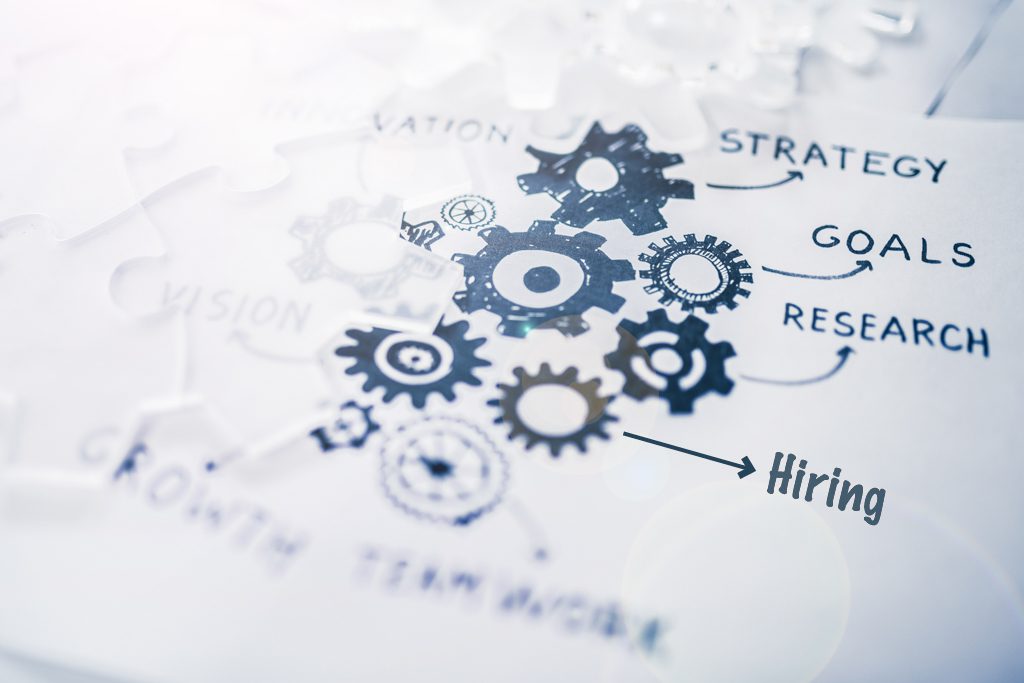 A business diagram on white paper with blue gears labelled "strategy," "goals" and "hiring."